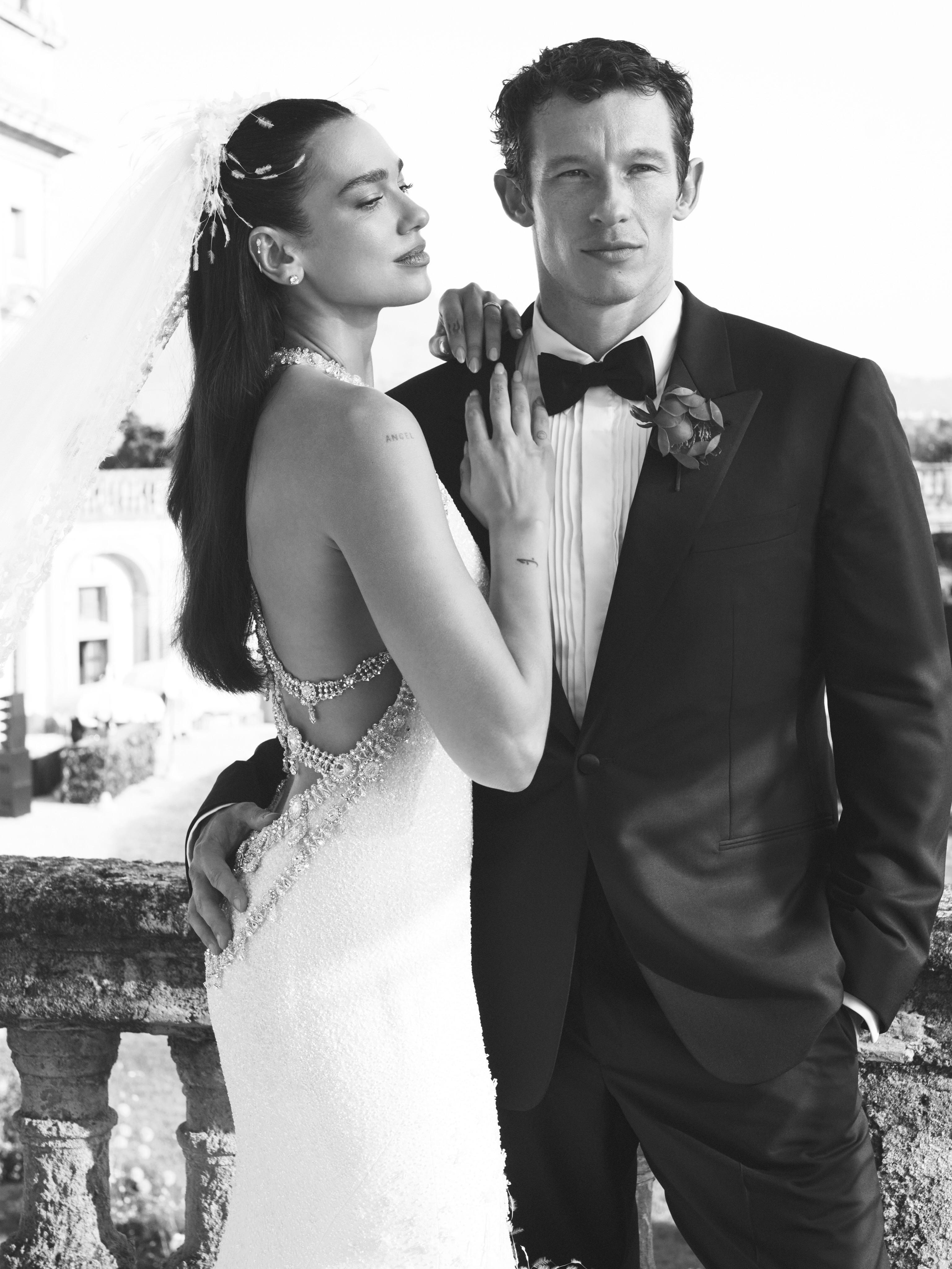 Black and white photo of Dua Lipa in a wedding gown with her arm around a man in a tuxedo.