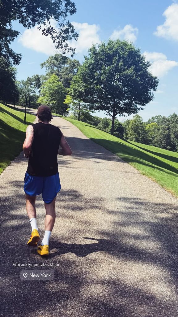 Brooklyn Beckham, in a black tank top and blue shorts, running uphill on a paved path surrounded by green grass and trees.