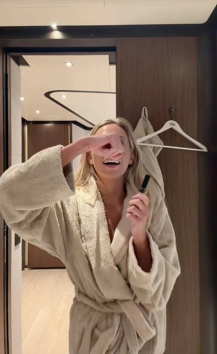 Alix Earle in a bathrobe applying makeup while holding a small black tube.