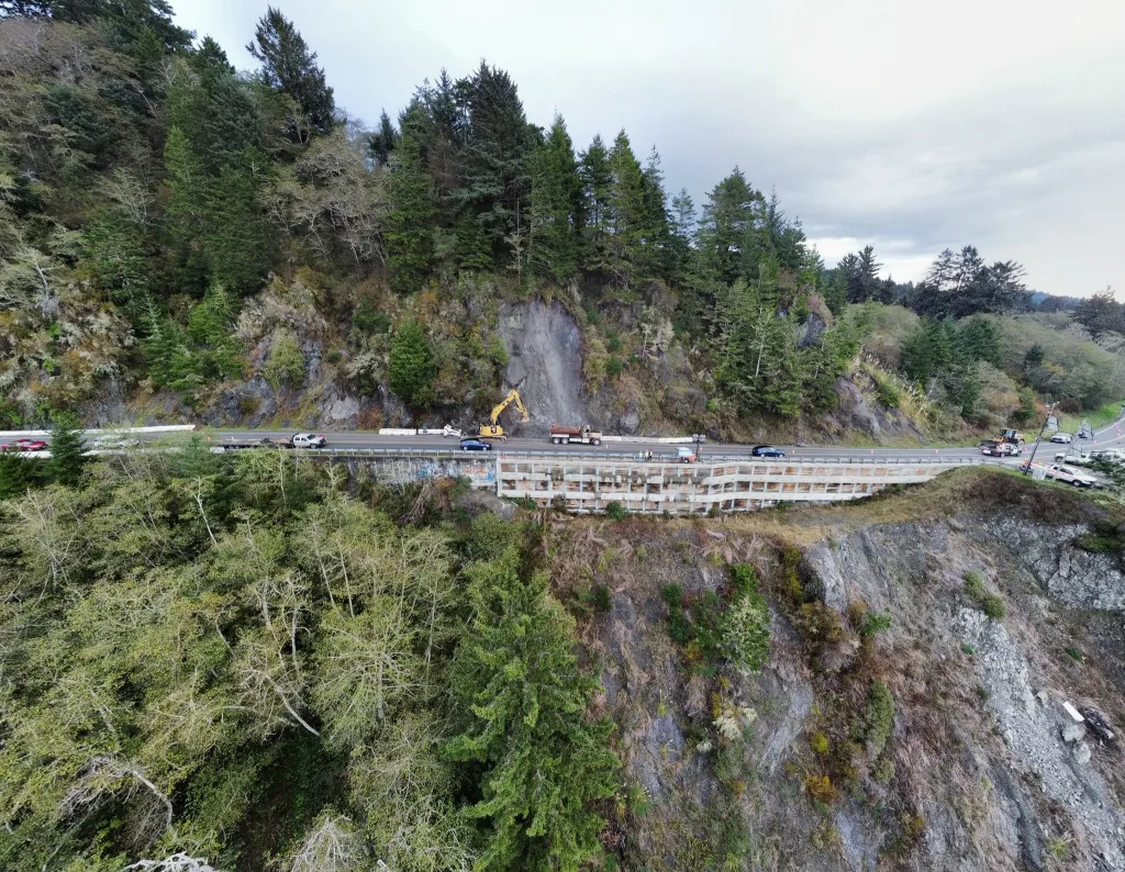 Construction vehicles on a highway next to a steep, tree-covered cliff.
