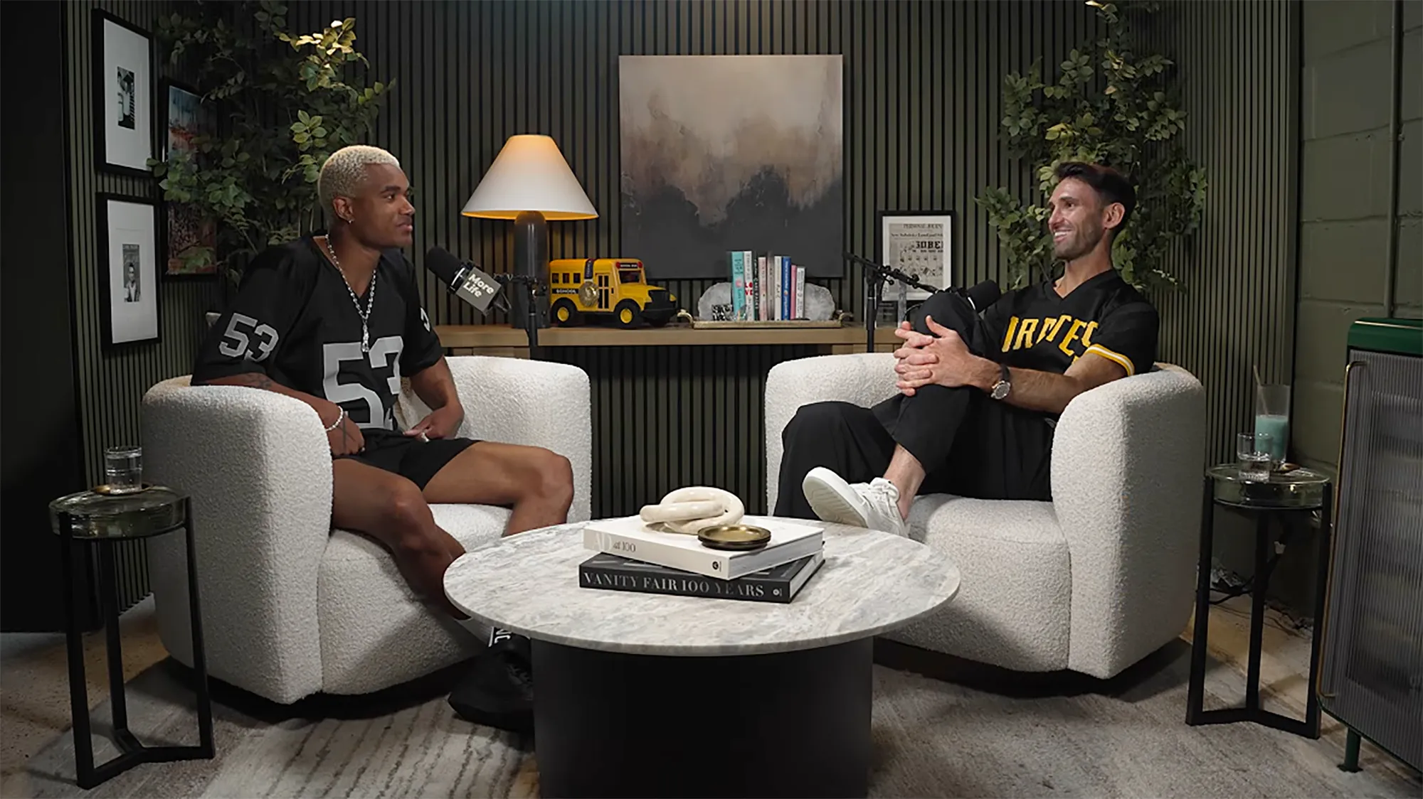 Two men in sports jerseys sit in armchairs, facing each other with microphones, in a podcast studio.