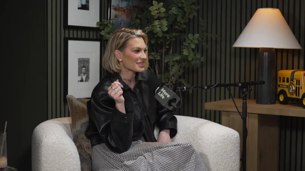 Woman in a black leather jacket and checkered pants sitting in a white armchair next to a microphone.