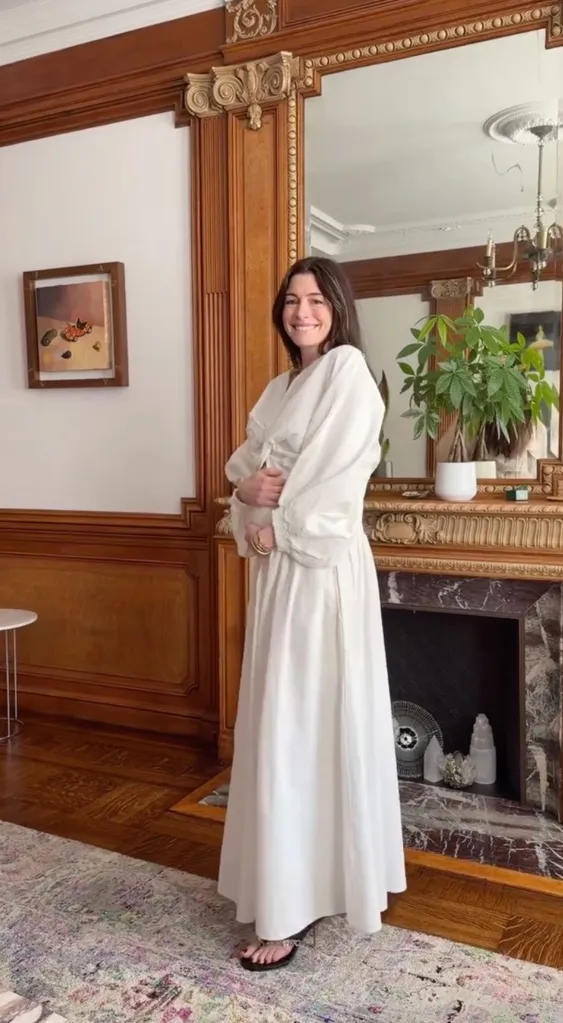 Anne Hathaway smiling in a white dress in a room with hardwood and a fireplace.