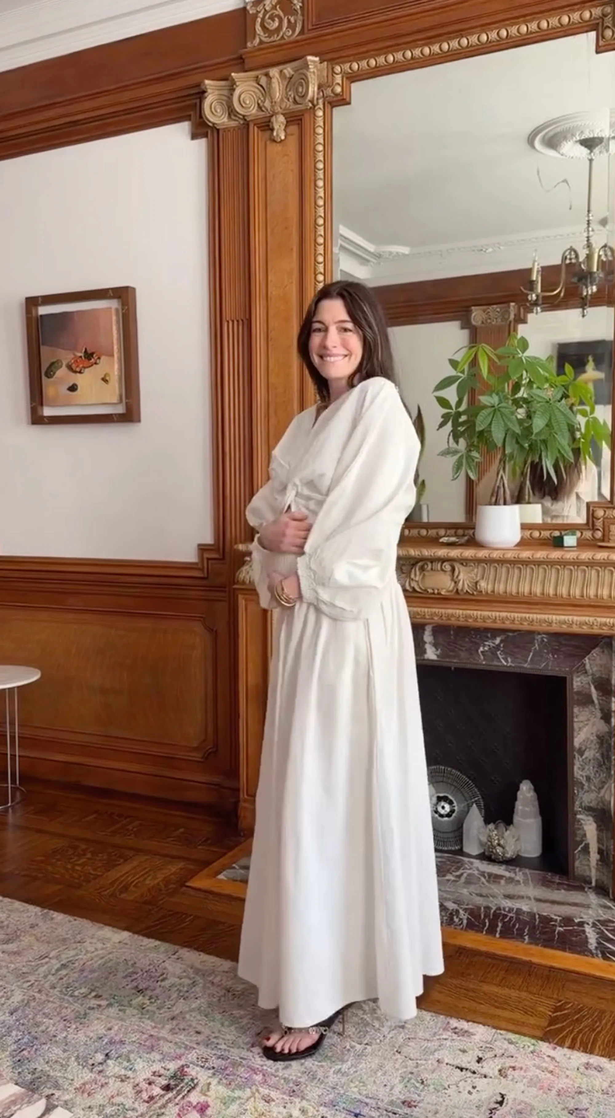 Anne Hathaway smiling and cradling her baby bump while wearing a long white dress.