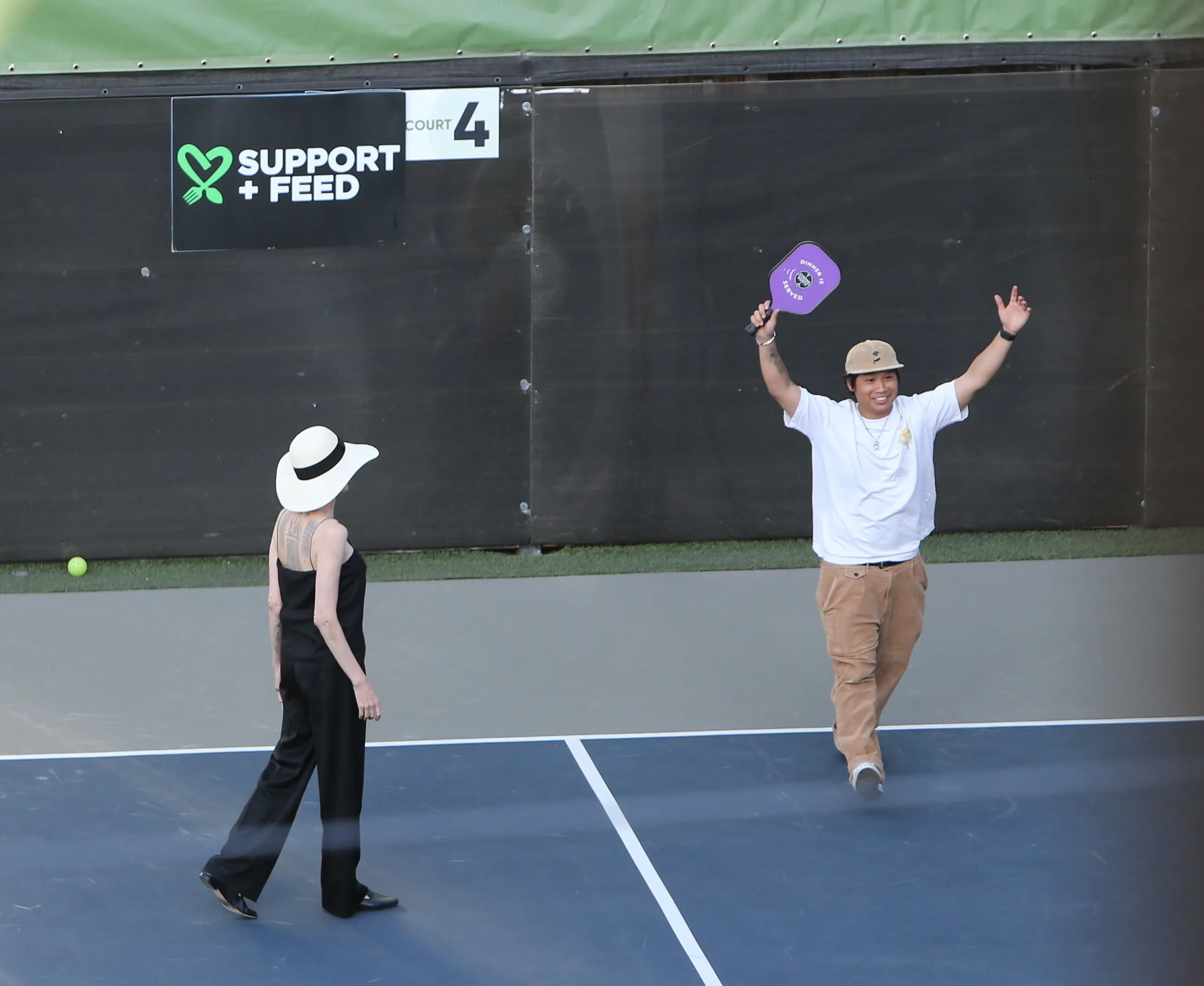 Angelina Jolie playing pickleball with her kids, dressed in a black outfit and white hat, stands on the court while a man celebrates with a purple paddle.