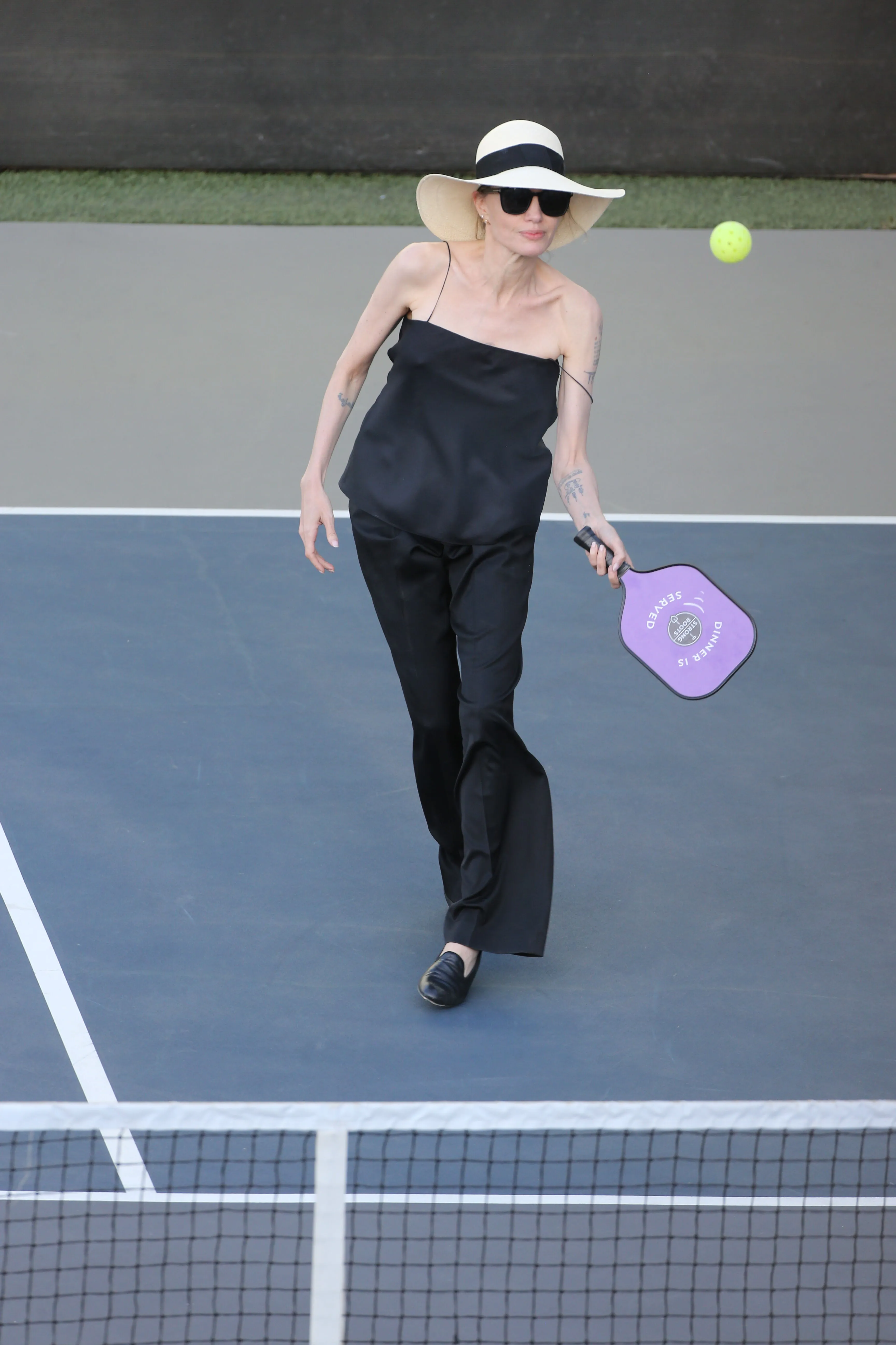 Angelina Jolie in a black top and pants, a sun hat, and sunglasses, holding a purple pickleball paddle on a pickleball court.