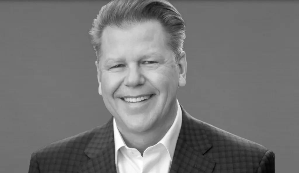 Portrait of Randy Kendrick, a man with neatly styled hair, wearing a white shirt and a checkered suit jacket, smiling at the camera.
