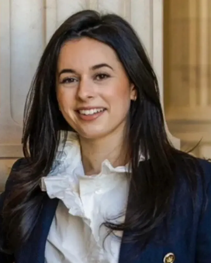 Yardena Wolf, with long dark hair, wearing a white ruffled blouse and a dark blazer.