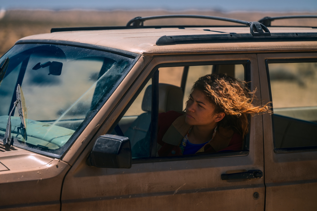 Zendaya as Rue in Euphoria Season 3, looking out the window of a dusty tan car with her curly hair blowing.