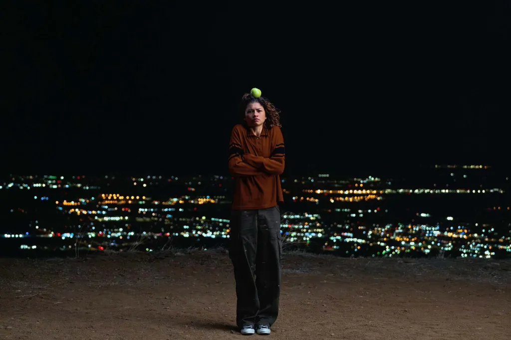 Zendaya as Rue Bennett with an apple on her head, arms crossed, standing on a hill at night with a city lights cityscape below.
