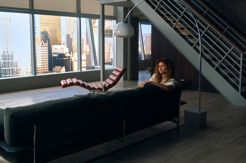 Zendaya as Rue in Euphoria Season 3, sitting on a couch in a modern apartment overlooking a city skyline.