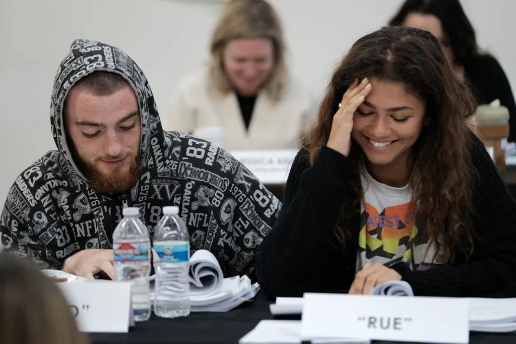 Angus Cloud in a Raiders hoodie looking at papers next to a smiling Zendaya, both sitting at a table with scripts.