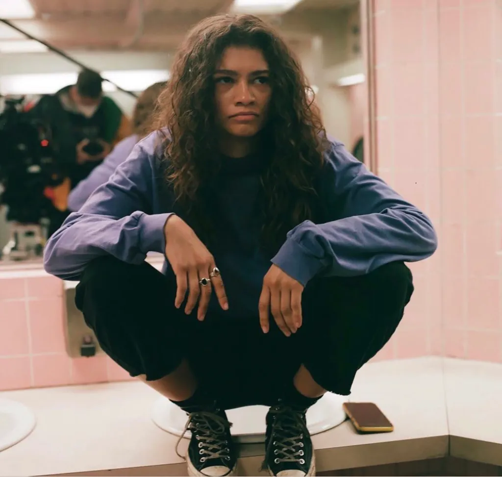 Zendaya as Rue on Euphoria, crouching in a public restroom with an indifferent expression.