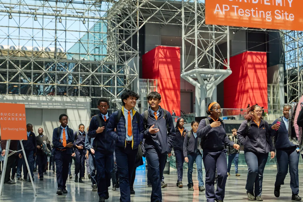 Students wearing navy uniforms with orange ties or shirts walk in a large hall with