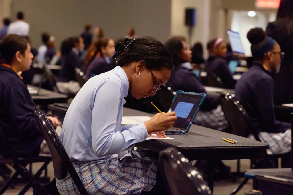 A student wearing glasses and a light blue shirt, writes on paper with a pencil in front of a laptop.
