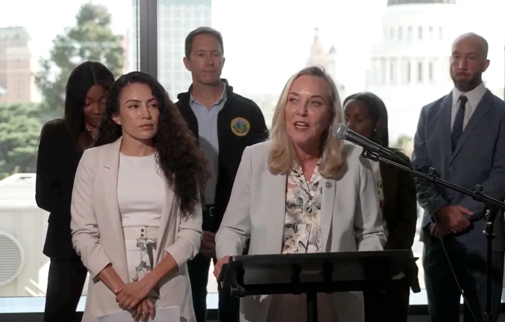 A woman speaking at a podium with a microphone, surrounded by other people, with a building visible through the window in the background.