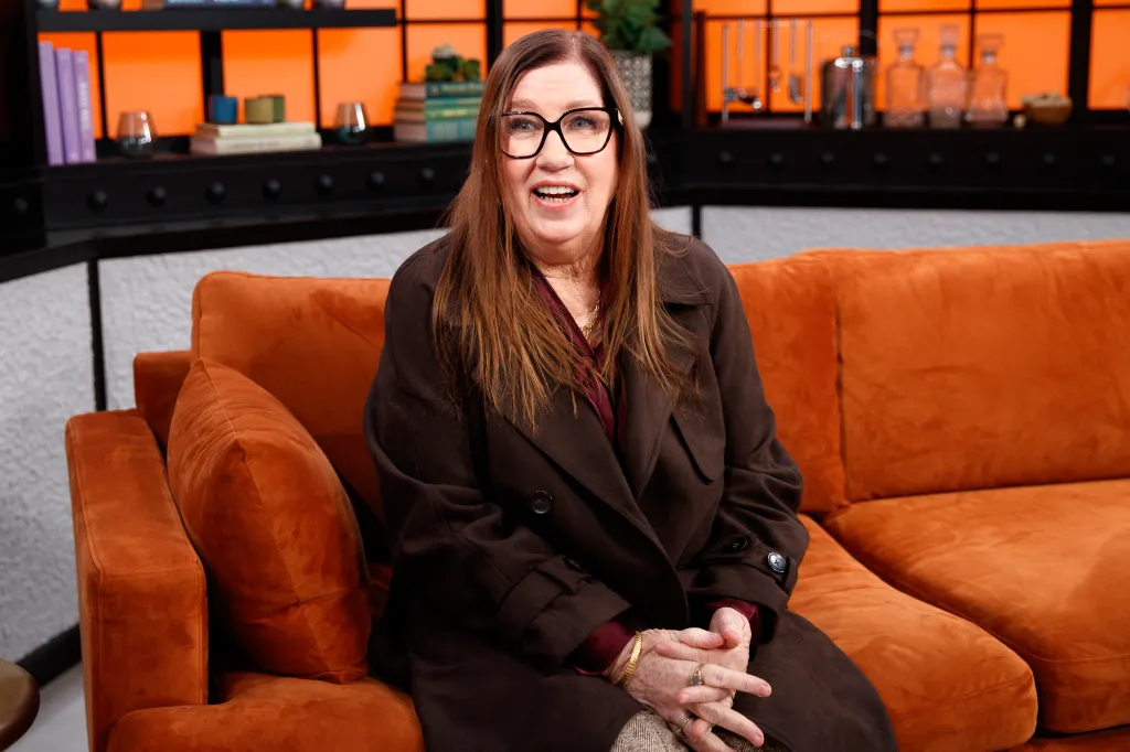 Jenny Slatten in a brown coat and glasses, seated on an orange couch.