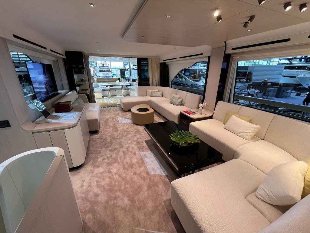 A modern yacht interior with light-colored sofas and a black coffee table, looking out onto a dock.