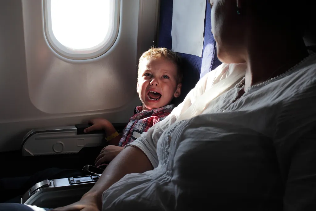 A child crying in an airplane seat next to an adult.