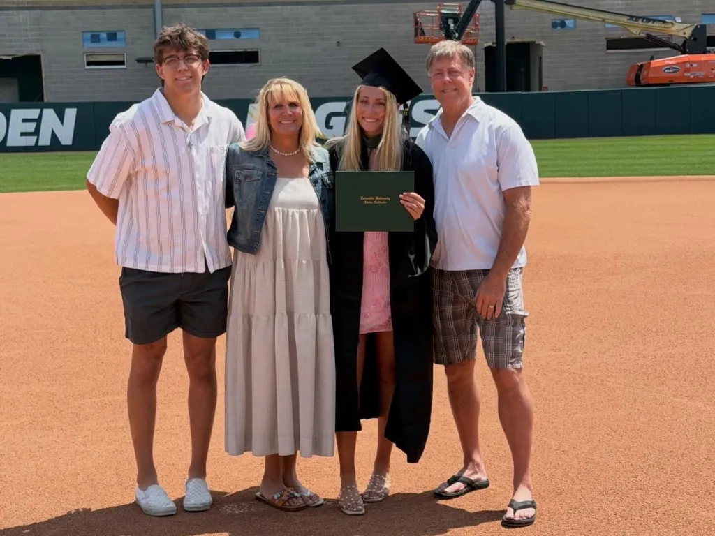 A young woman in a graduation cap and gown holding a diploma stands with her family on a baseball field.
