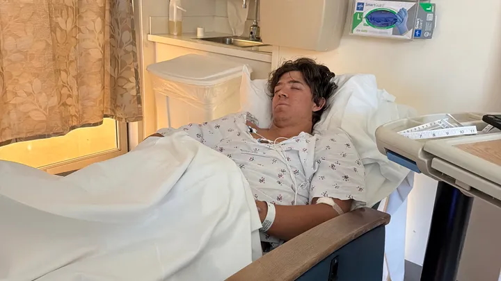 A young man lies in a hospital bed with wires attached to his chest.