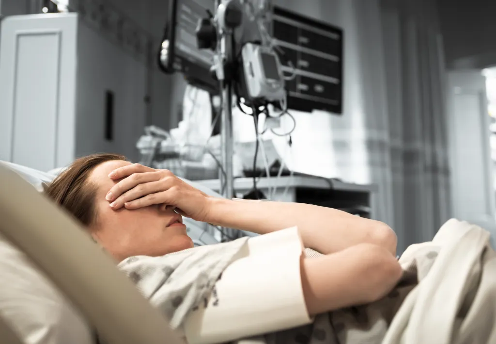 A woman lies in a hospital bed with her hand over her eyes, appearing to be in pain.