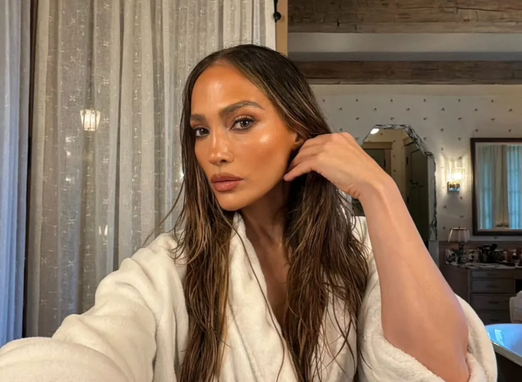 Jennifer Lopez in a white robe with wet hair, touching her ear.