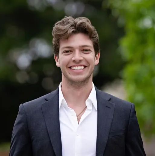 A young man with light brown, wavy hair smiles, wearing a dark suit jacket over an unbuttoned white shirt with a gold chain visible.