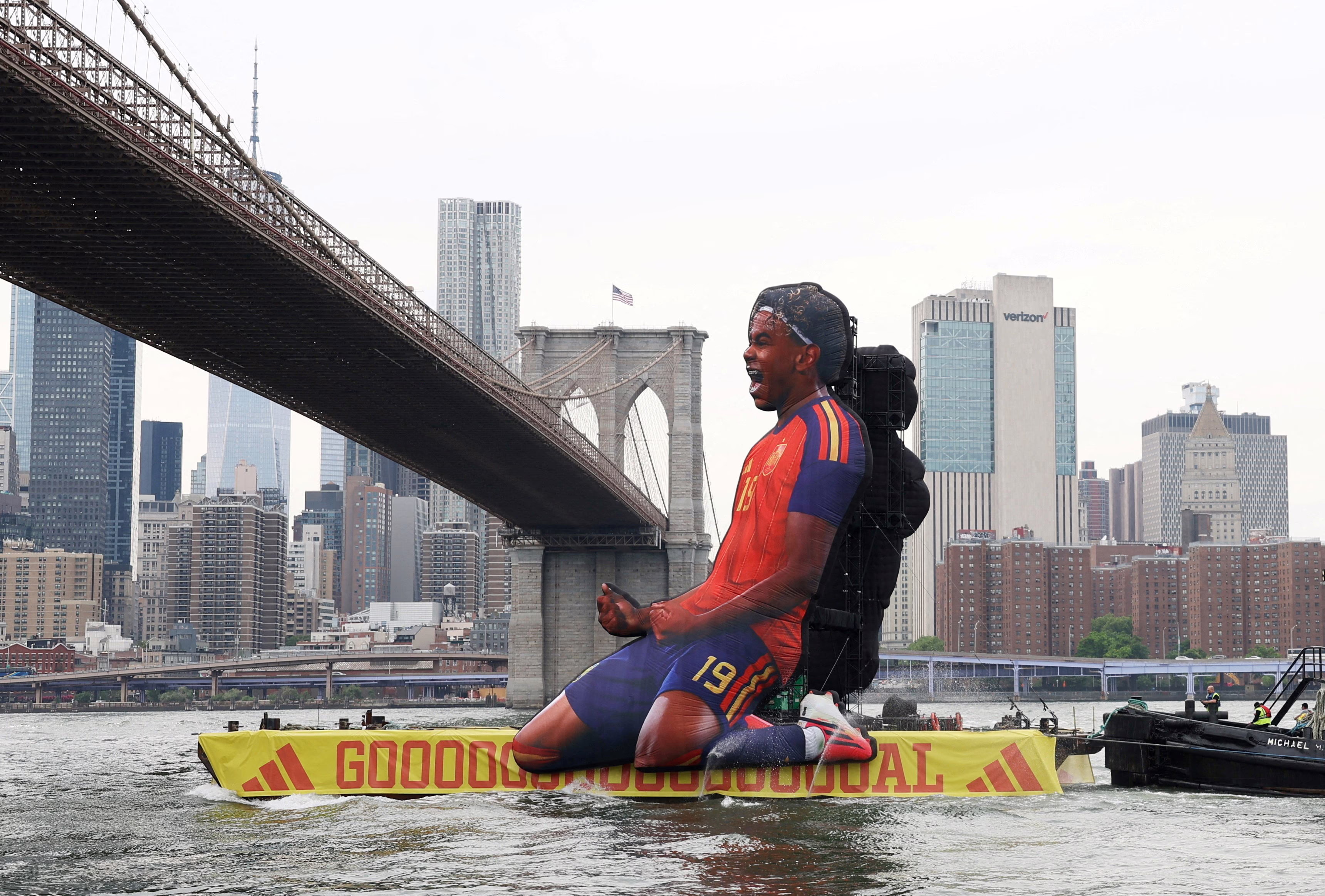 A barge, decorated with a tall advertising billboard shaped like a soccer player sliding on their knees, passes beneath the Brooklyn Bridge.