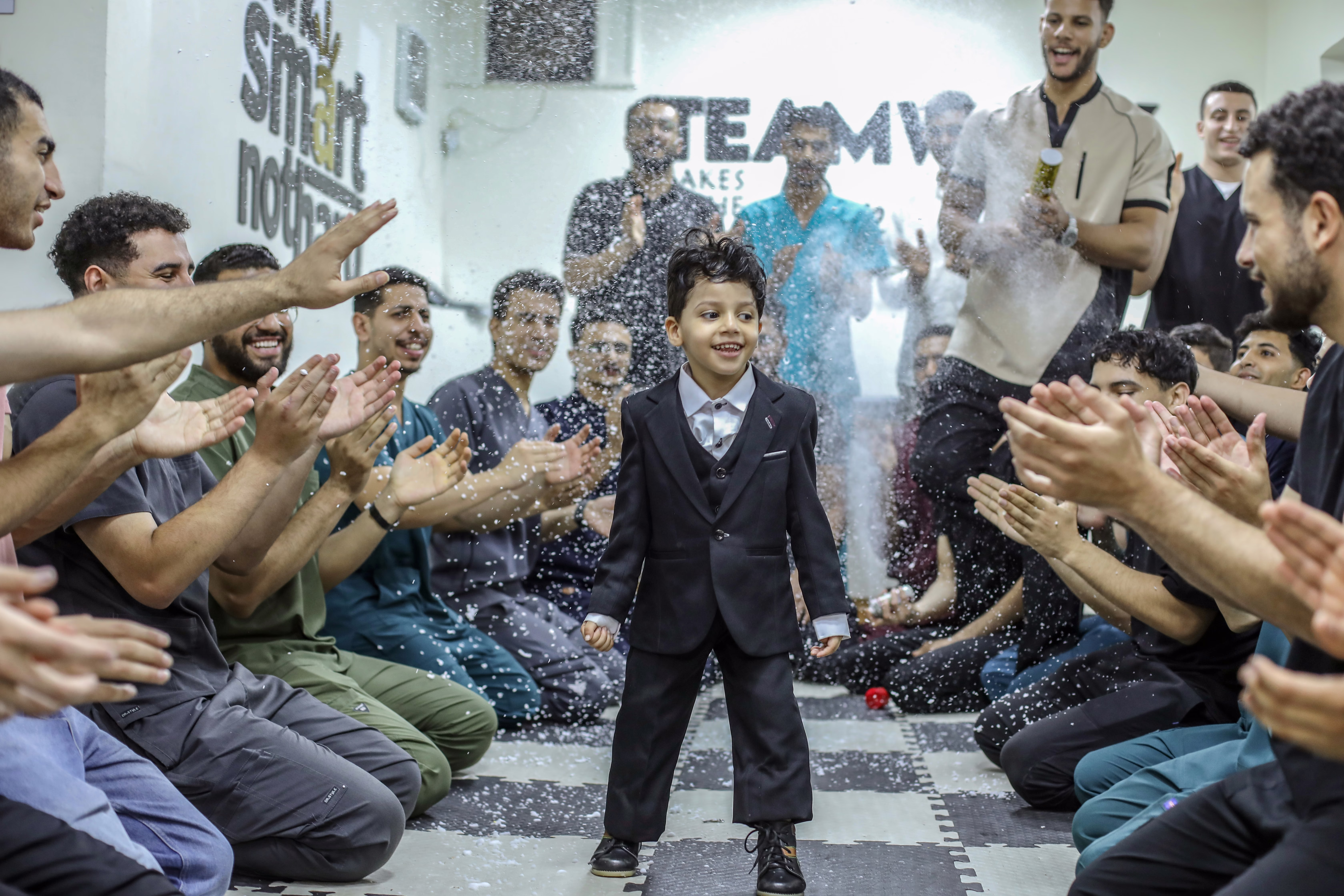 A young boy in a suit smiles as he is surrounded by cheering staff members at a rehabilitation center. 