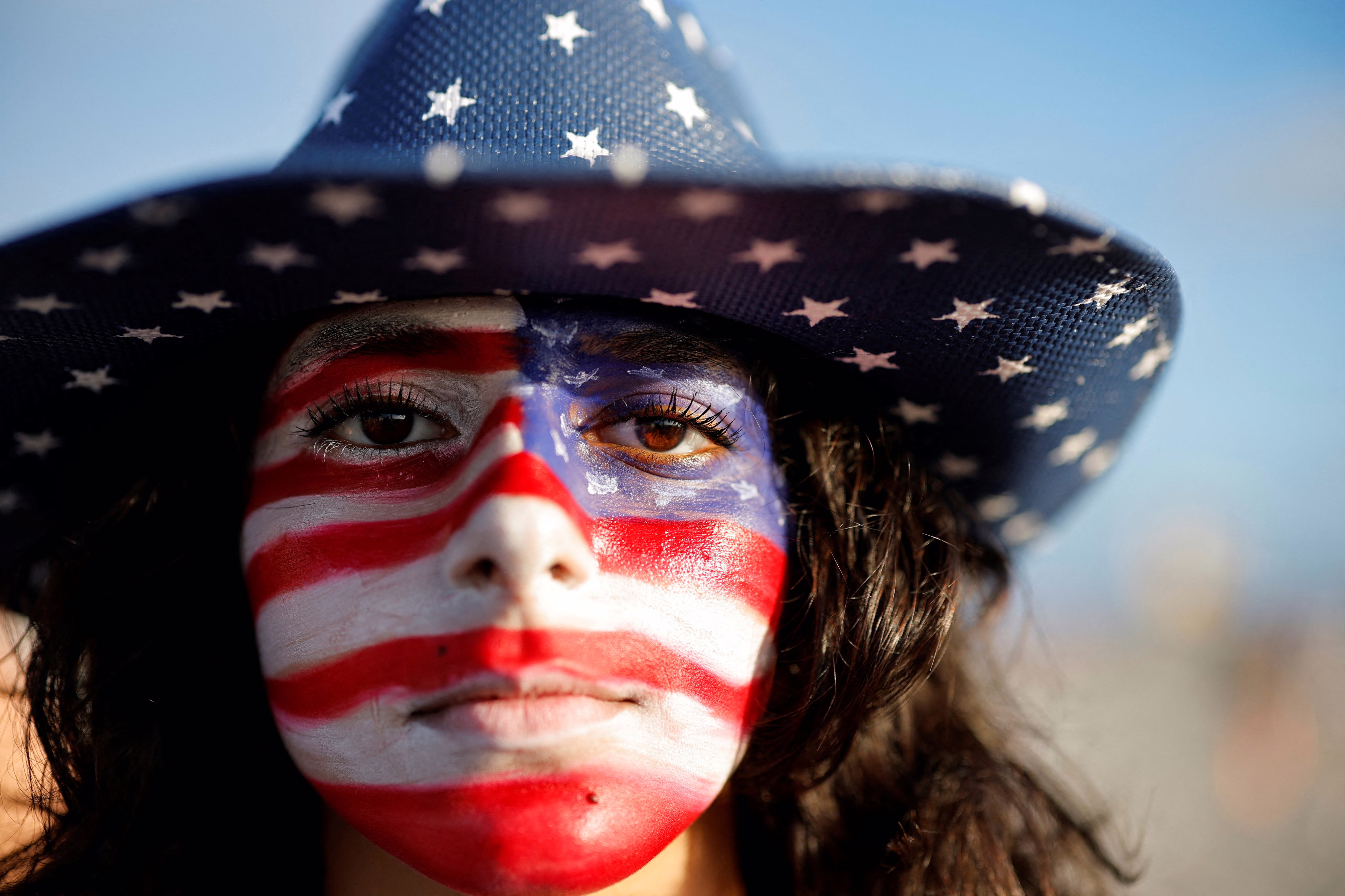 A woman poses with her face painted in the colors of the U.S. flag, wearing a blue cowboy hat with white stars.