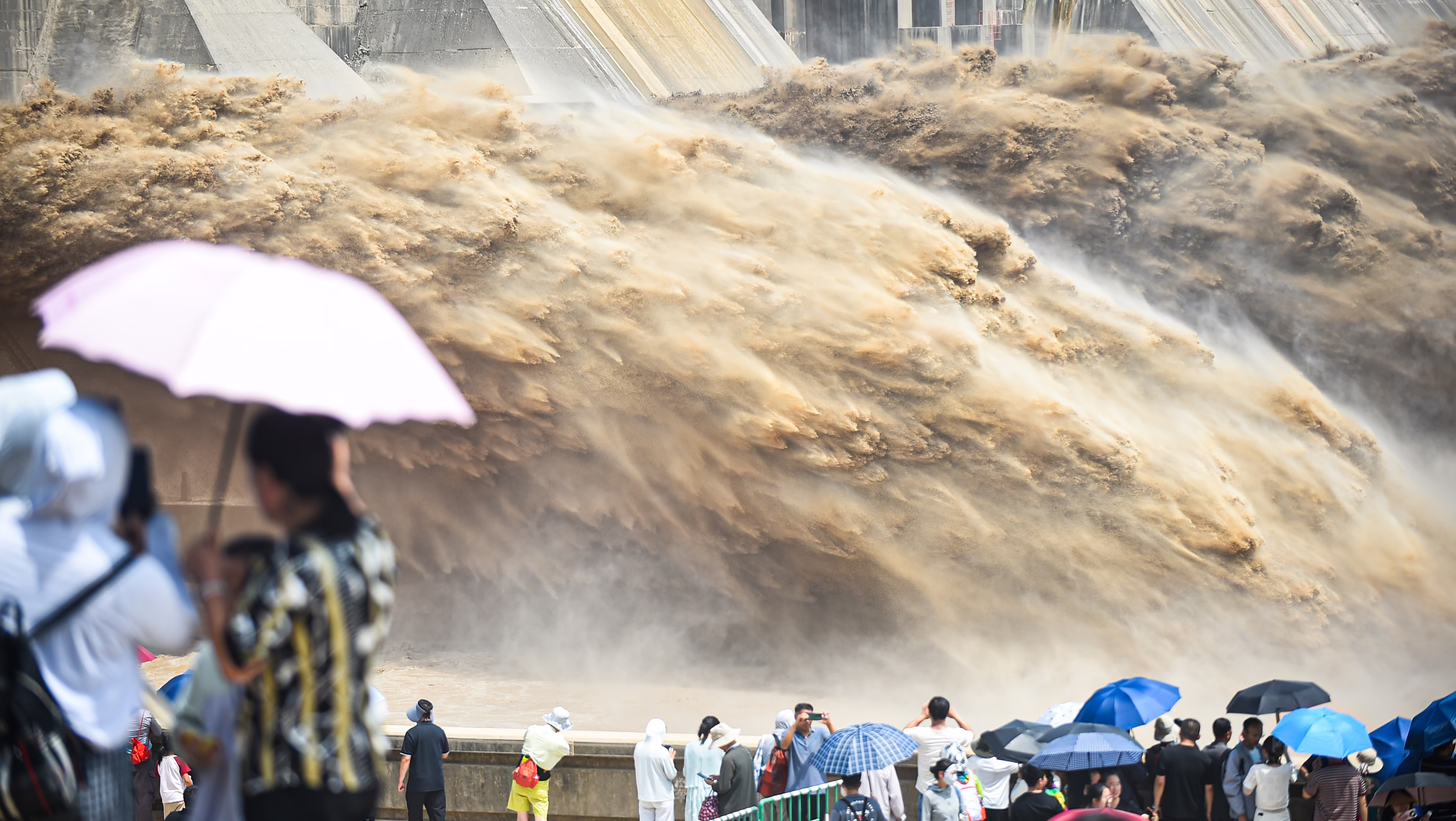 Tourists watch huge jets of muddy water flowing out of the bottom of a tall dam.