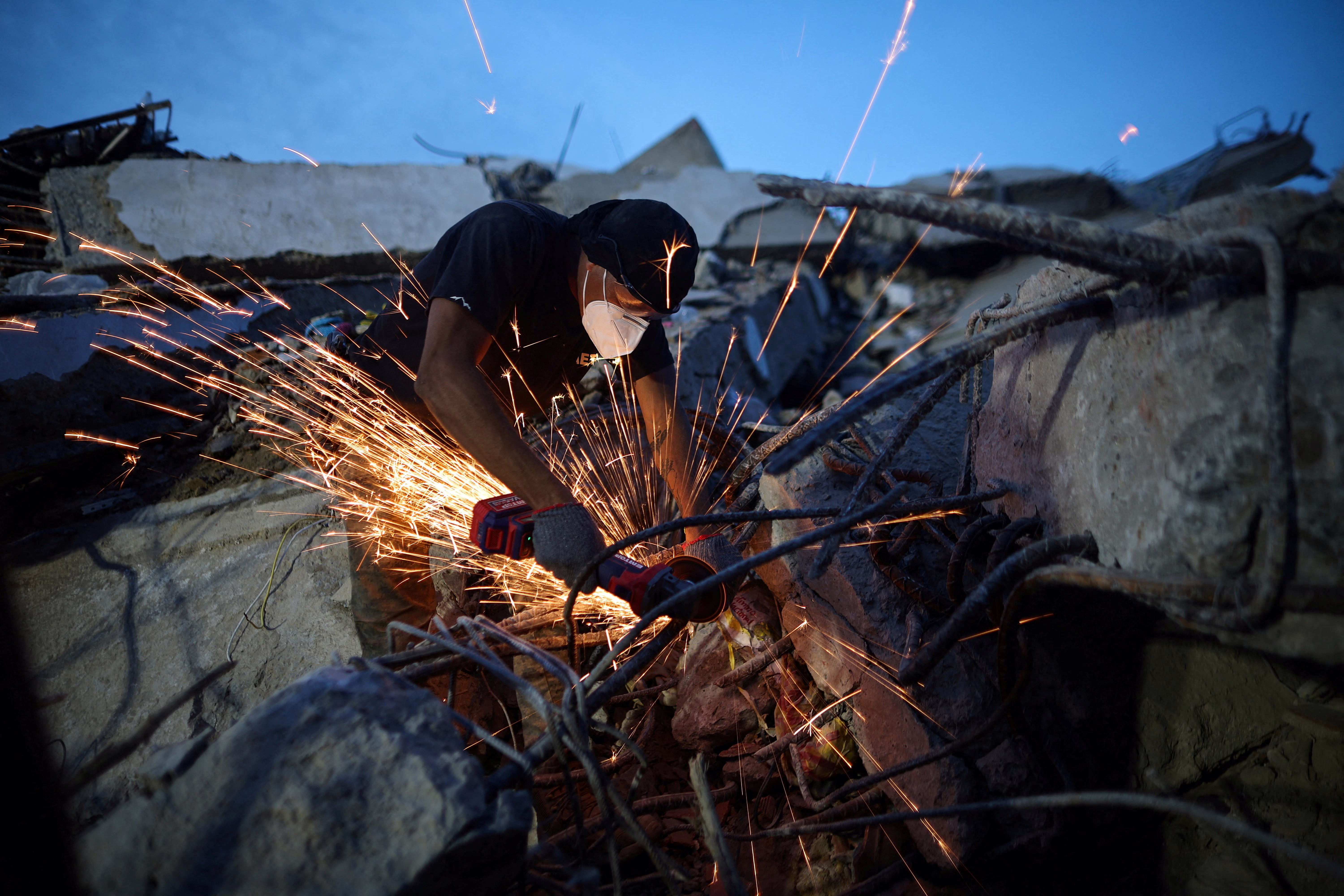 A man uses an angle grinder at the site of a collapsed building, sending a shower of sparks out over building debris.