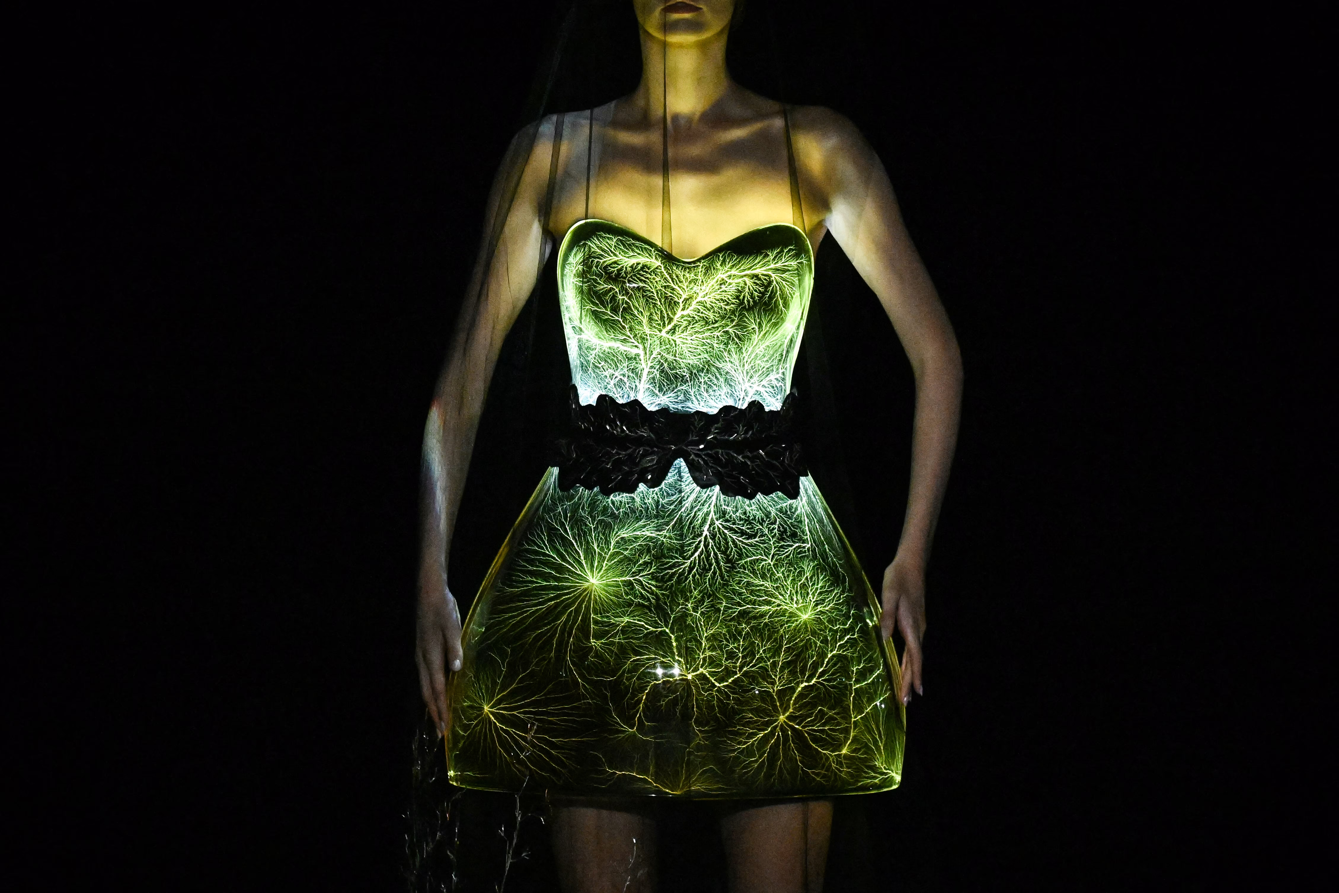 A model poses, wearing a dress that appears to be made of glass or plastic, displaying branching cracks, lit from within.