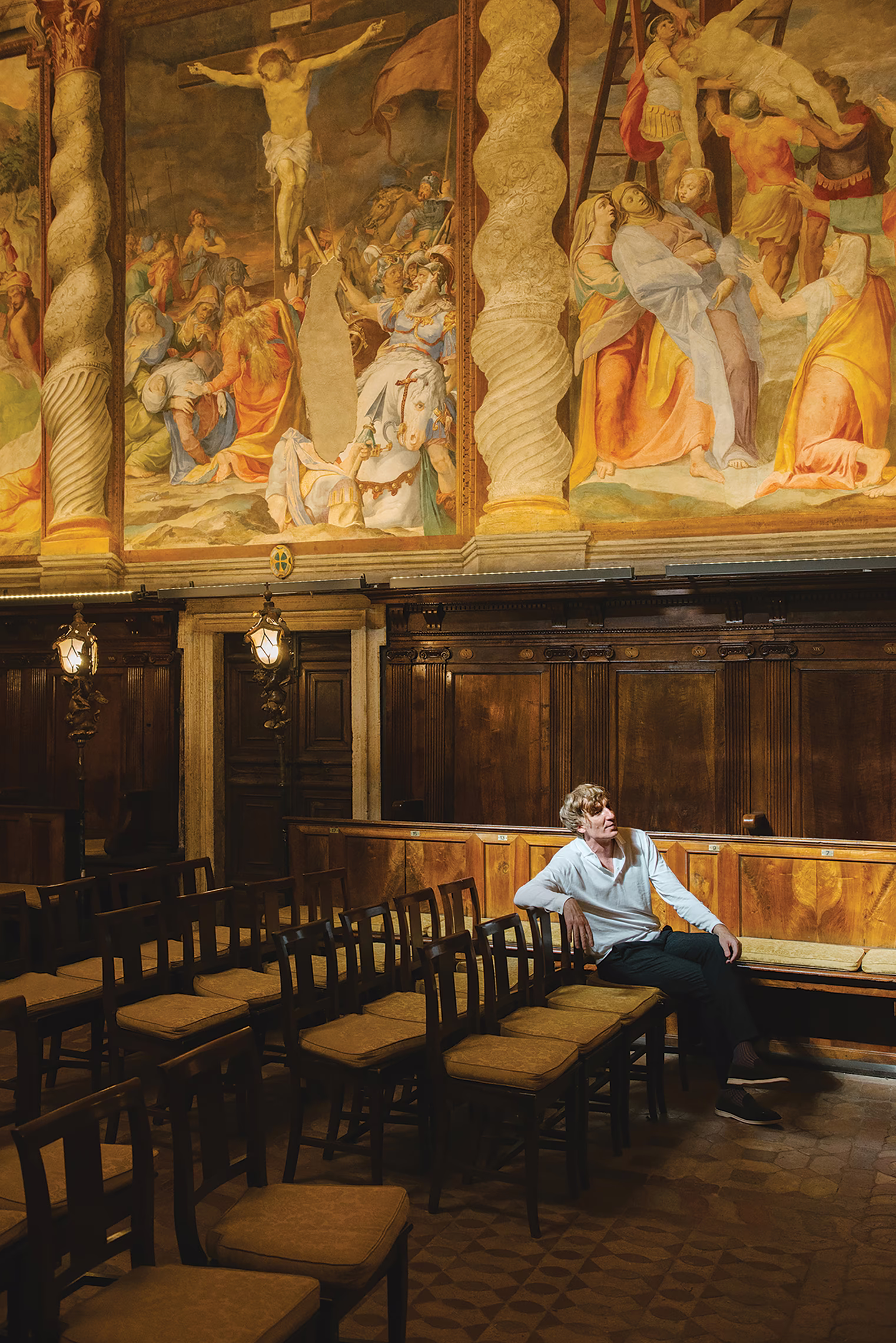 photo of a man sitting in front row of chairs in room covered in paintings
