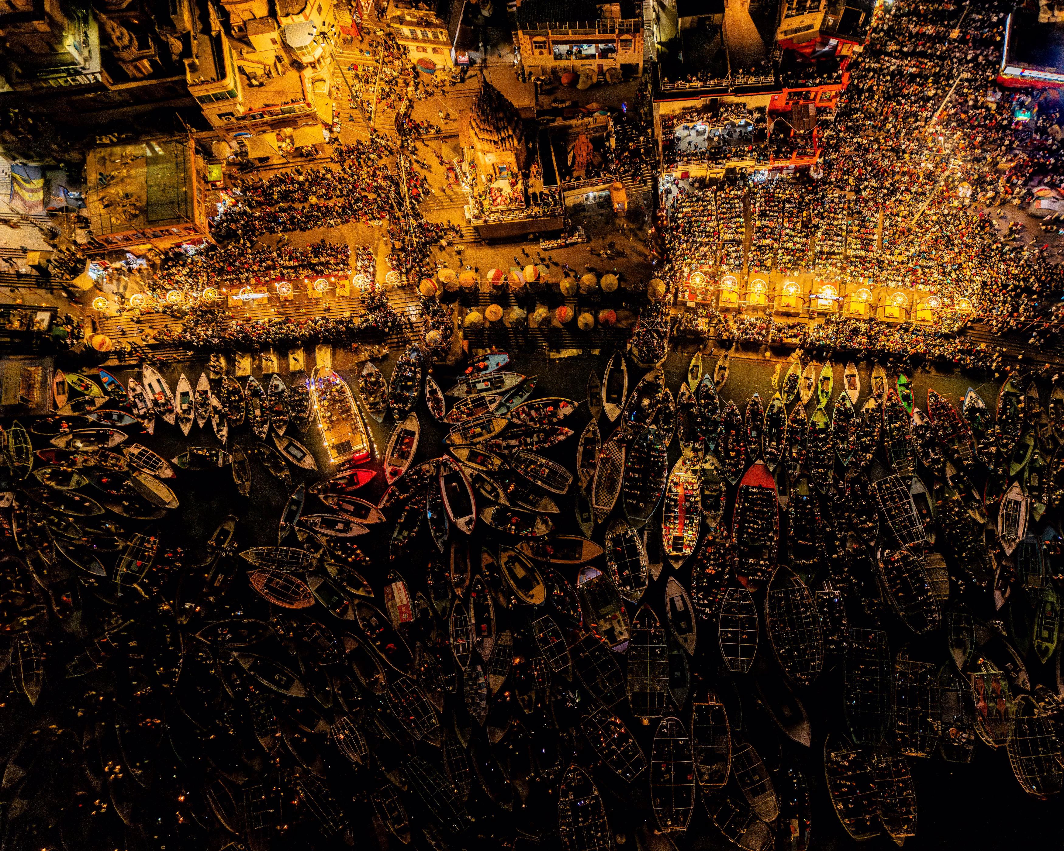 An aerial view of a riverbank packed with people, on shore and in small boats near the shore, seen at night, in India