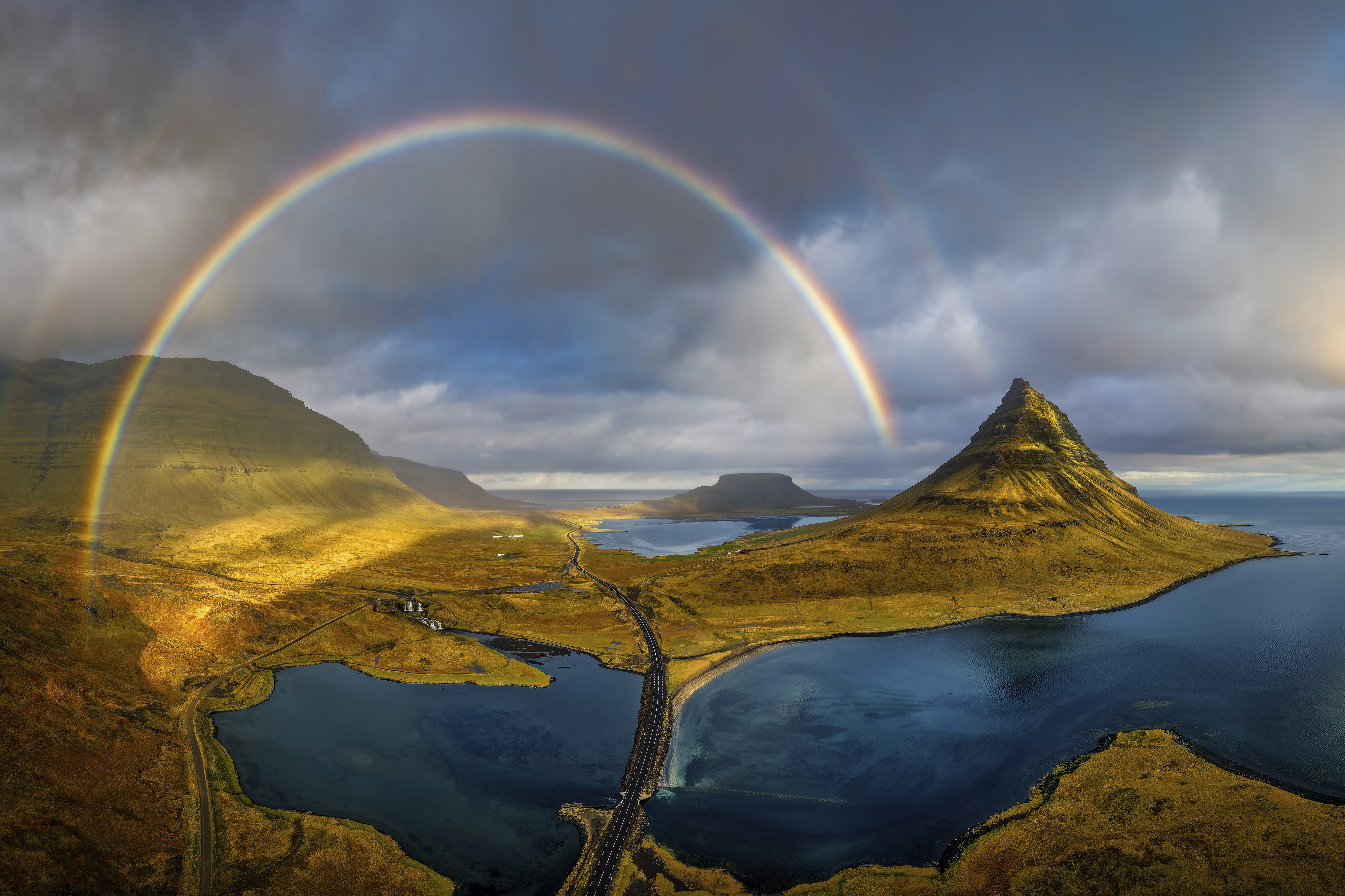 An aerial panorama of coastal hills and mountains in Iceland, covered in greenery, beneath a rainbow