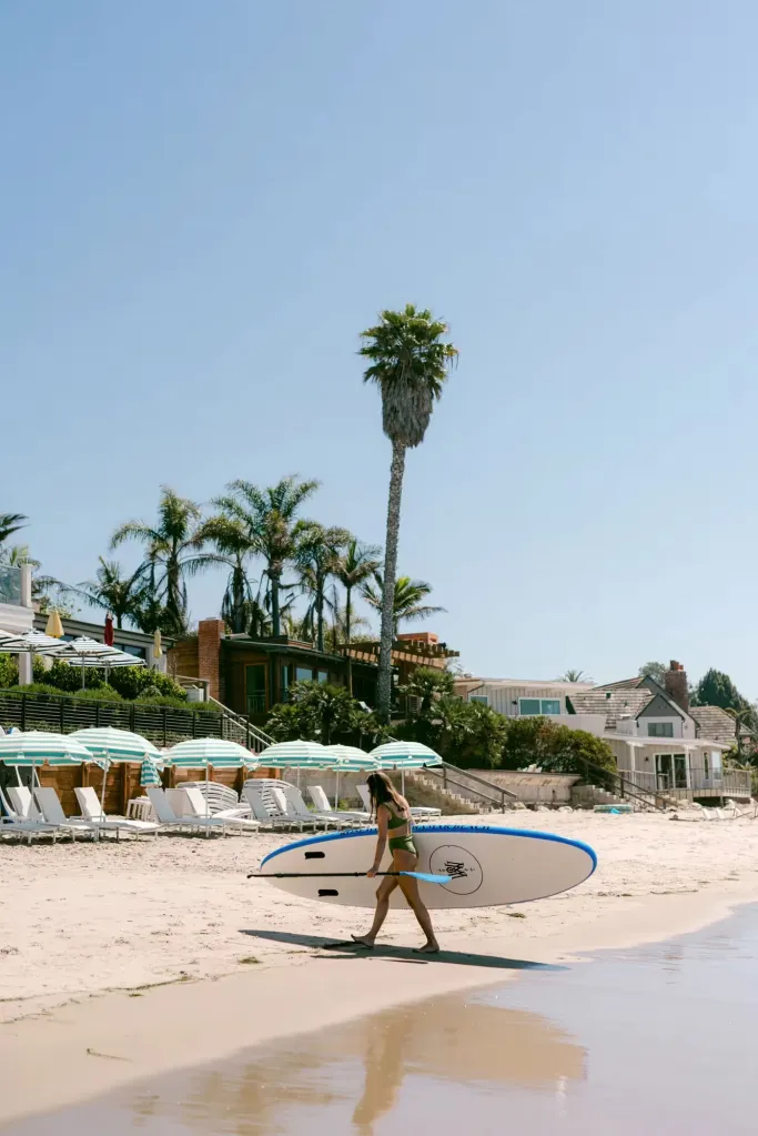 A woman in a green bikini carrying a paddleboard on the beach in front of Rosewood Miramar Beach.