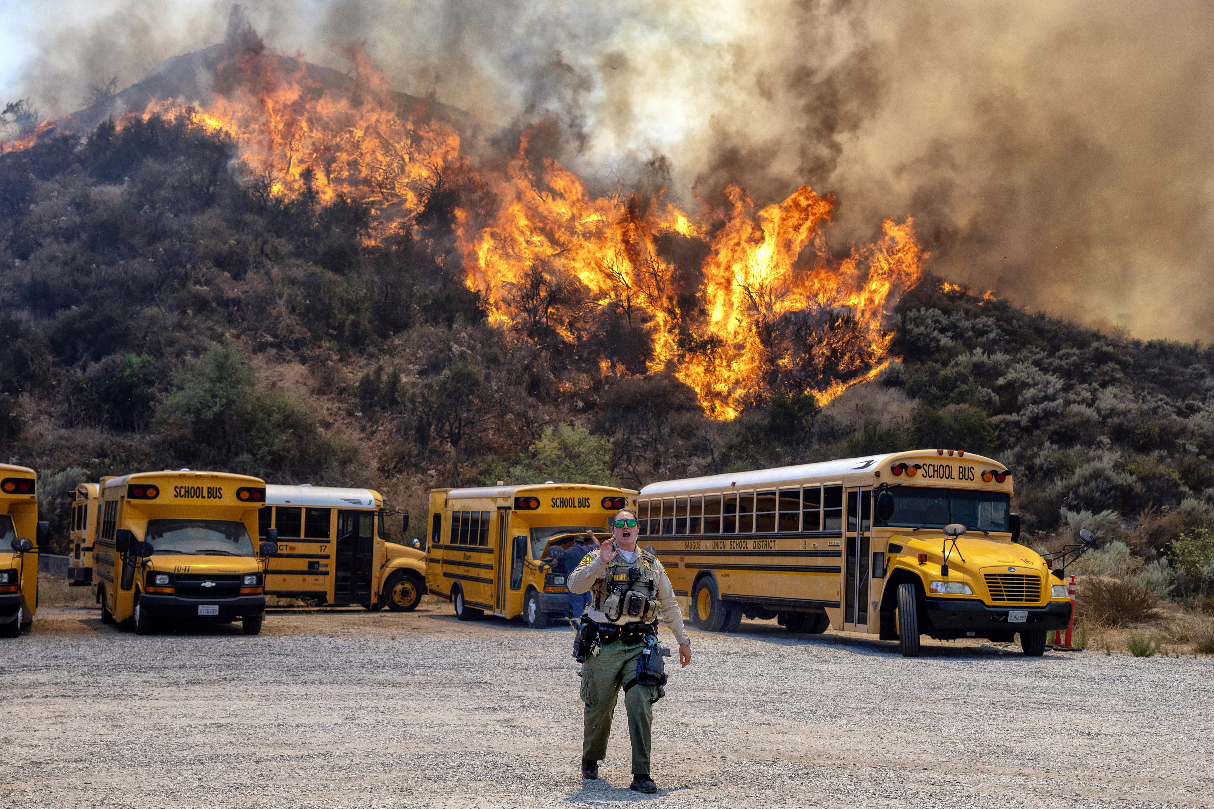 A brush fire burns on a hillside above several school buses parked in a lot.