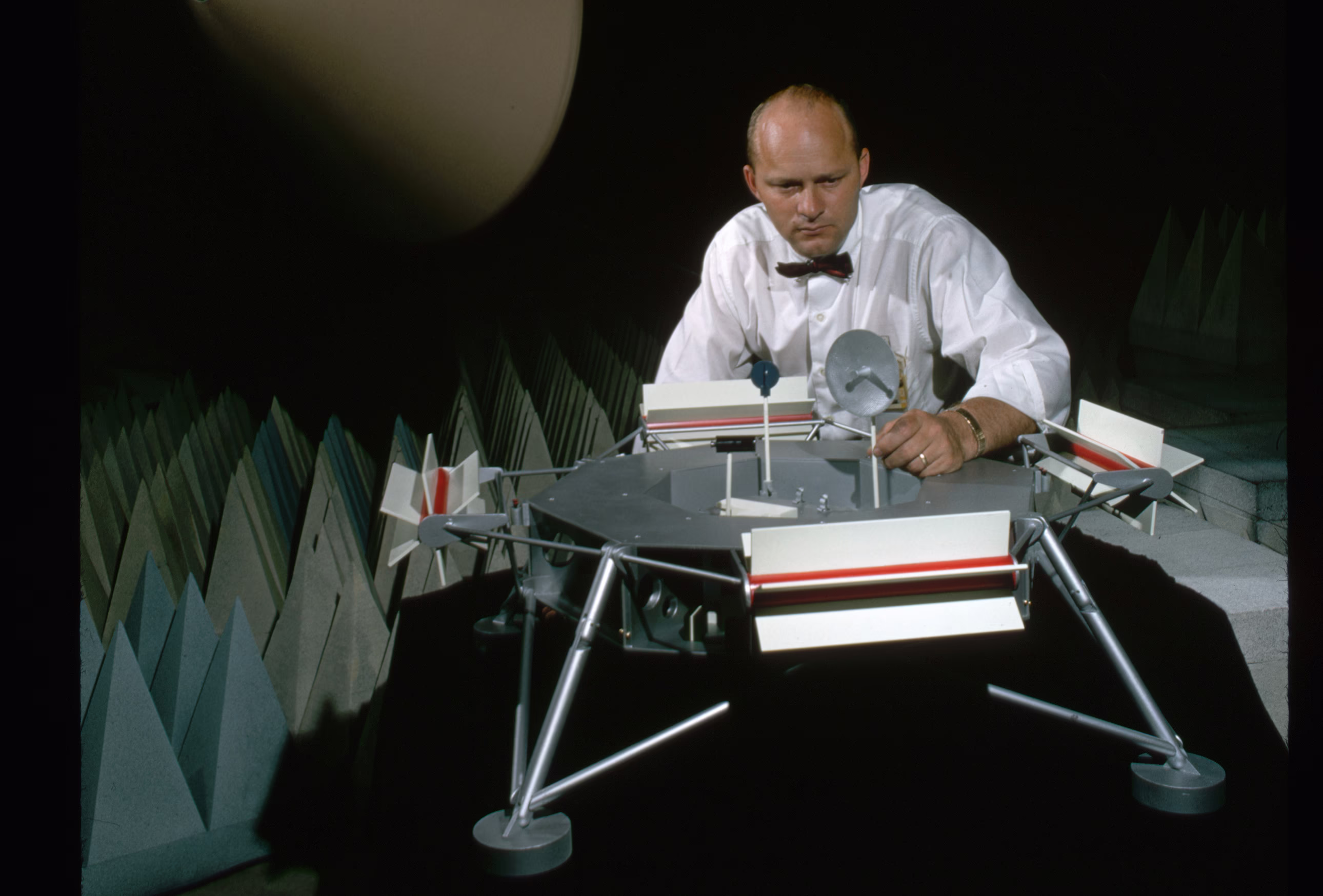 A man sets up a model of the Mars Viking spacecraft in what appears to be a sound-dampening room with conical baffles behind him.