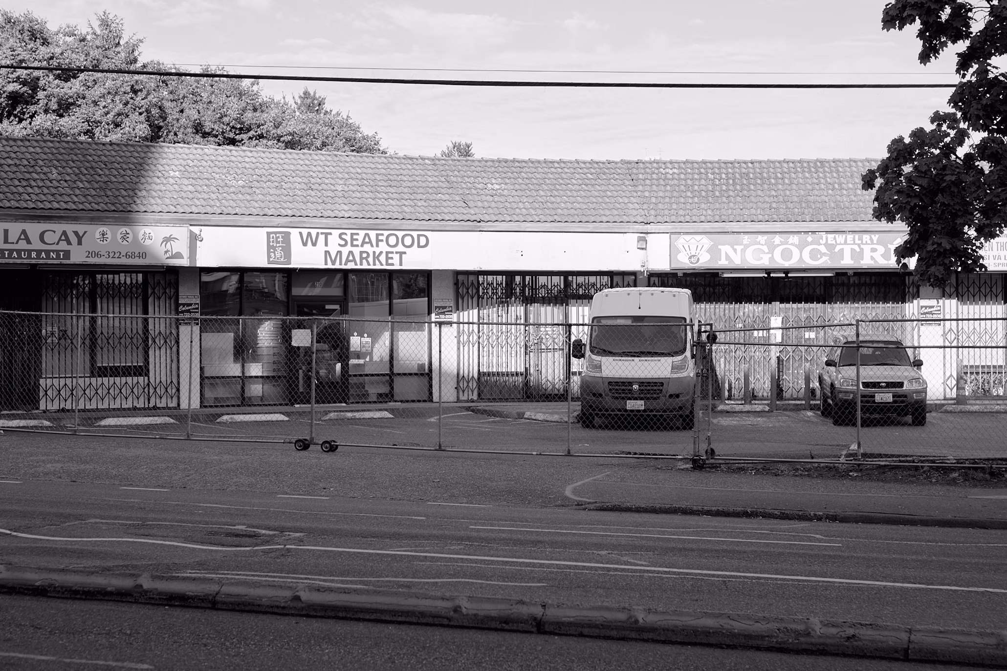 black-and-white photo of strip center shops with chain-link gates across parking lot