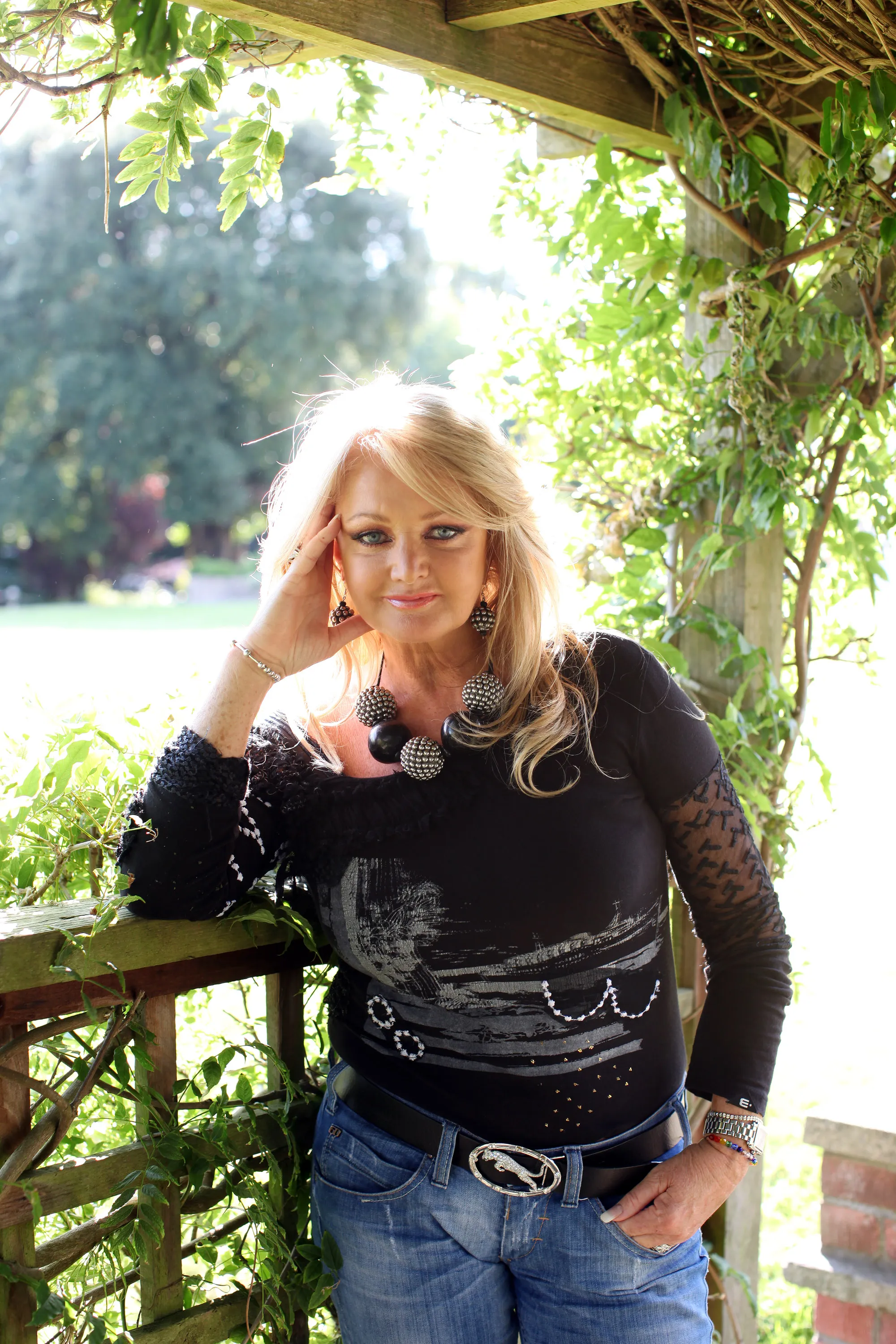 Bonnie Tyler leaning against a wooden trellis outdoors.