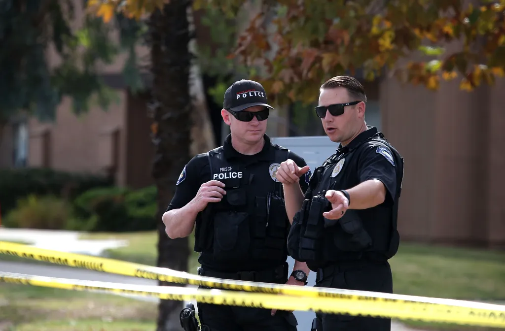 Two police officers in tactical gear and sunglasses standing behind yellow police tape.