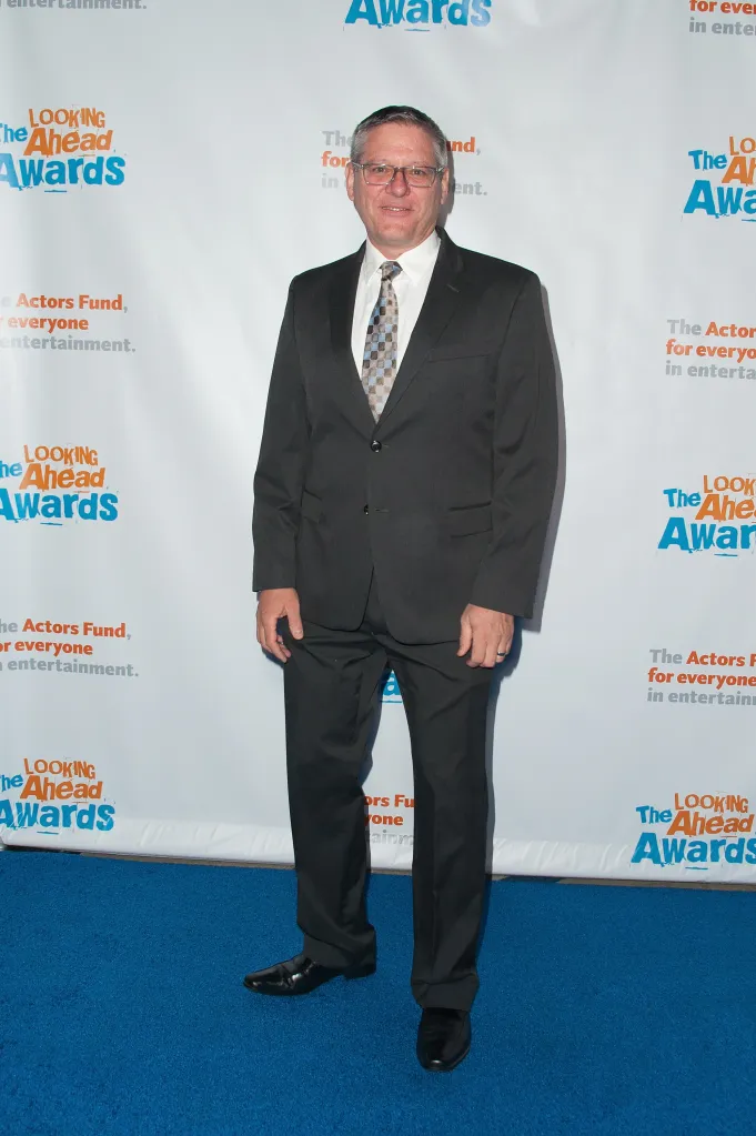 Radames Pera smiling and wearing a suit and patterned tie on a blue carpet in front of a white backdrop with 