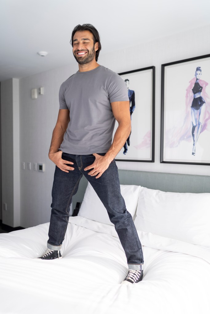 Sam Asghari, in a grey T-shirt and blue jeans, stands on a white bed, smiling with his eyes closed.