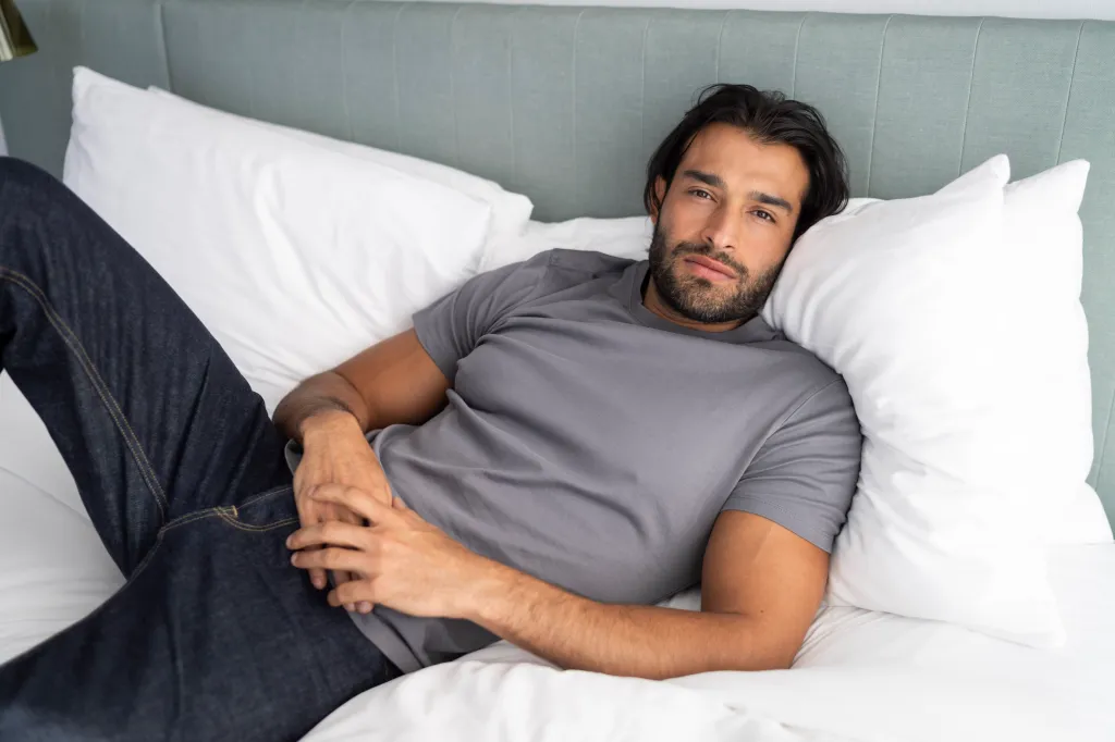 Britney Spears' ex-husband, Sam Asghari, reclines on a white bed with a light grey headboard. He wears a grey T-shirt and dark blue jeans.