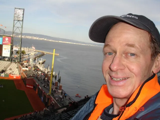 Steven Bassett smiling while wearing a baseball cap and orange jacket at a ballpark overlooking the bay.