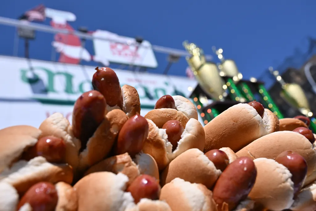 Hot dogs in buns ready for the Nathan's Fourth of July Hot Dog contest, with a blurred banner and trophies in the background.