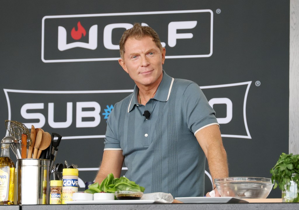 Bobby Flay standing behind a counter with ingredients and kitchen utensils, in front of a dark gray wall with 