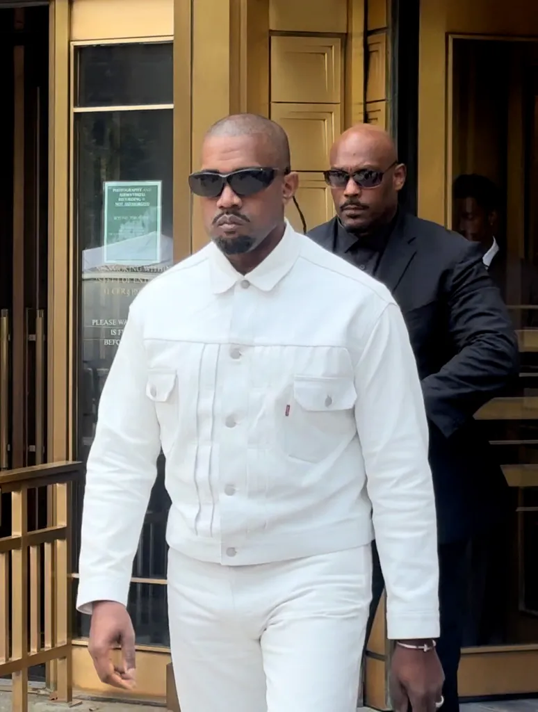 Kanye West, wearing a white denim outfit and sunglasses, leaving a courthouse with a bald security guard behind him.
